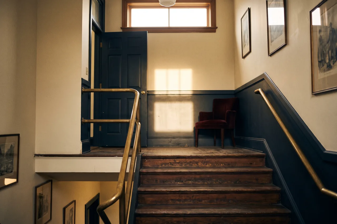 The wooden stairwell of the Aldous Block with a brass handrail and morning light through the transom