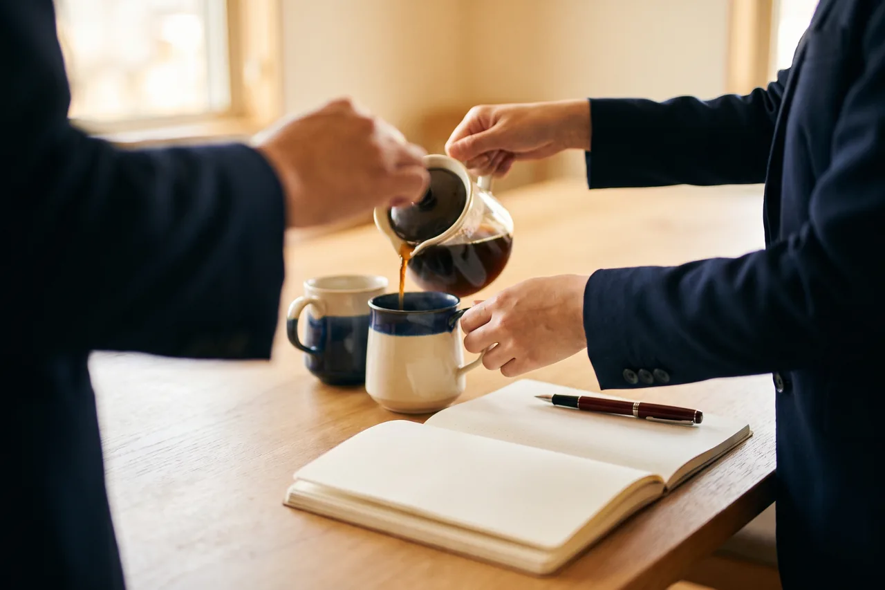 A woman's hands pouring coffee into two cups on a meeting table beside a notepad