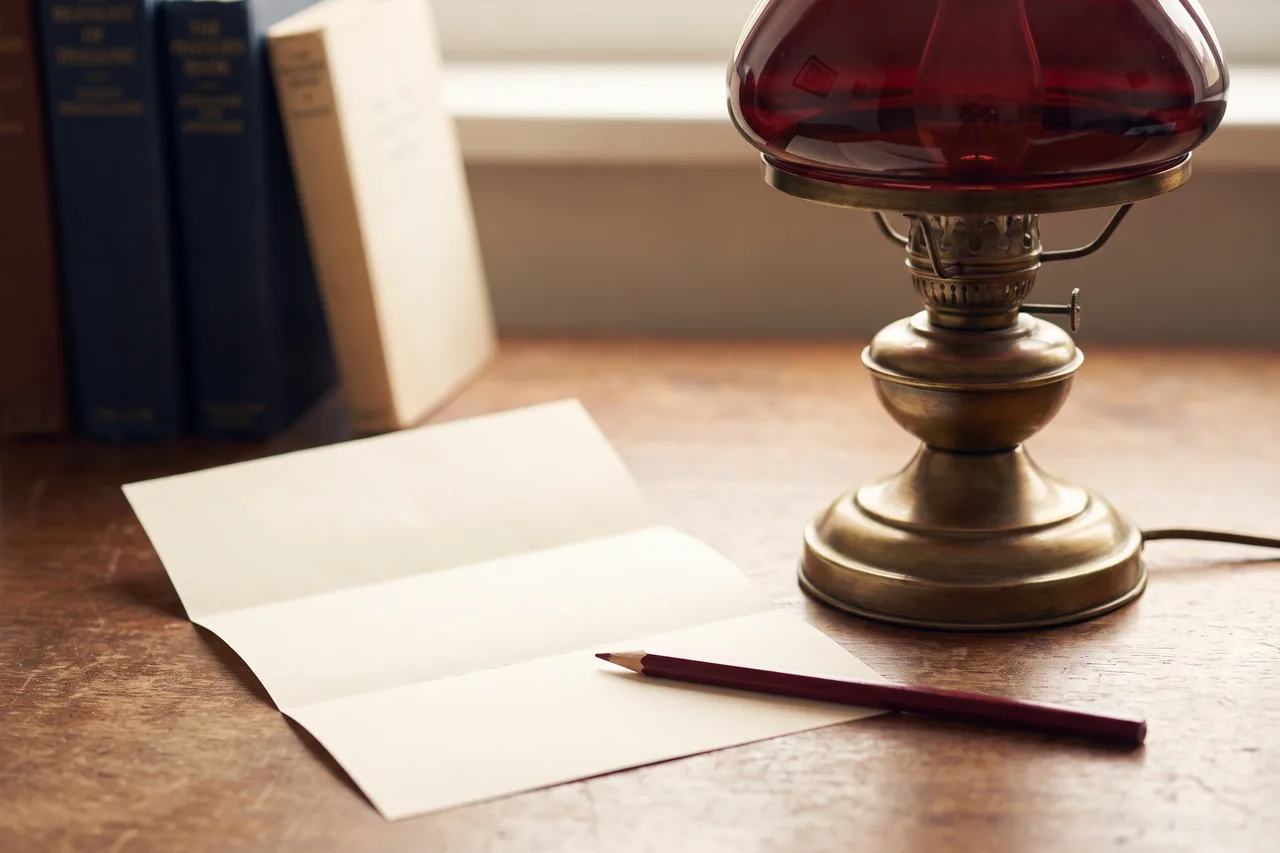 A folded letter on a desk beside an oxblood pencil and a green-shaded reading lamp
